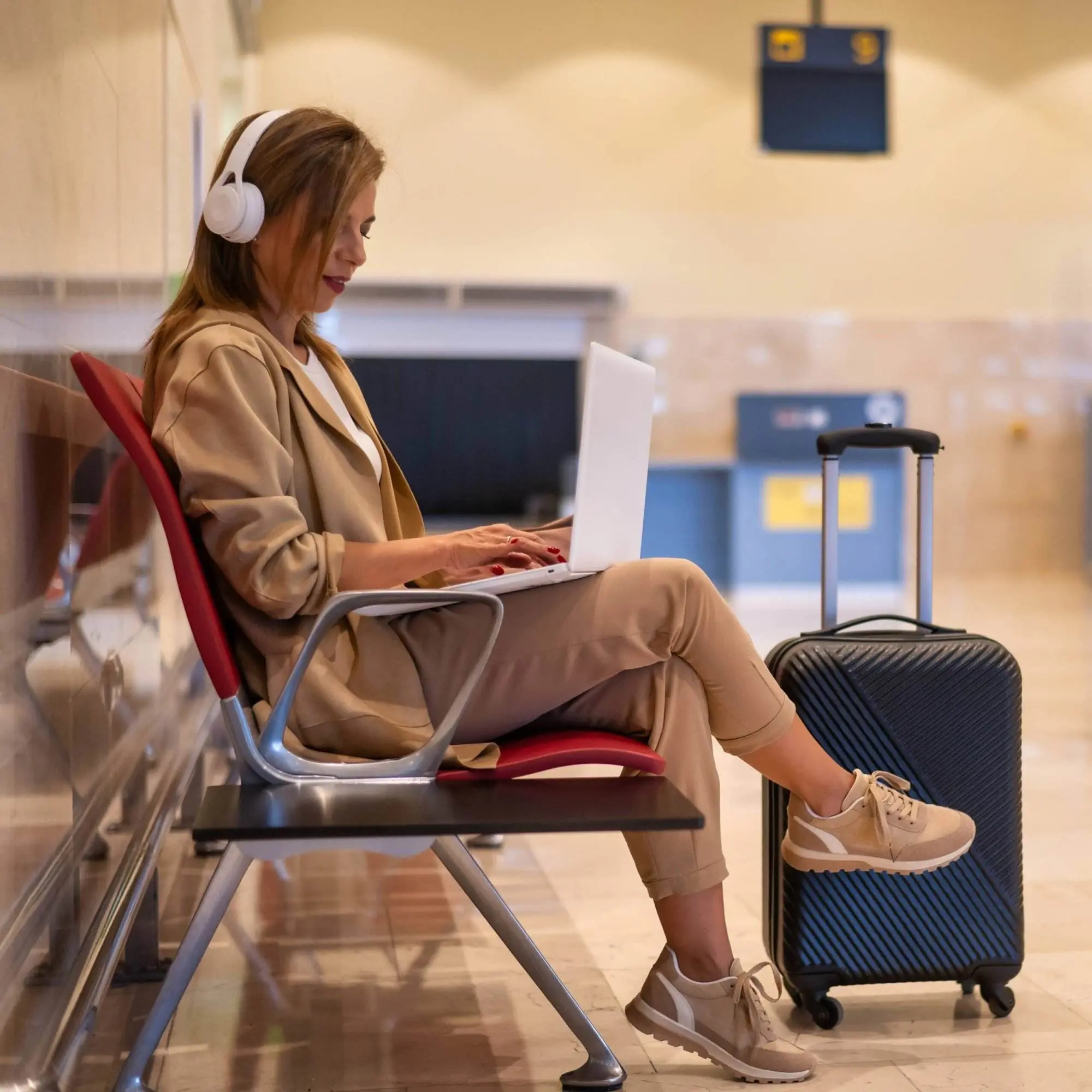 A lady is sitting in the airport wearing over-the-ear headphones while typing on her laptop. Her luggage is next to her.