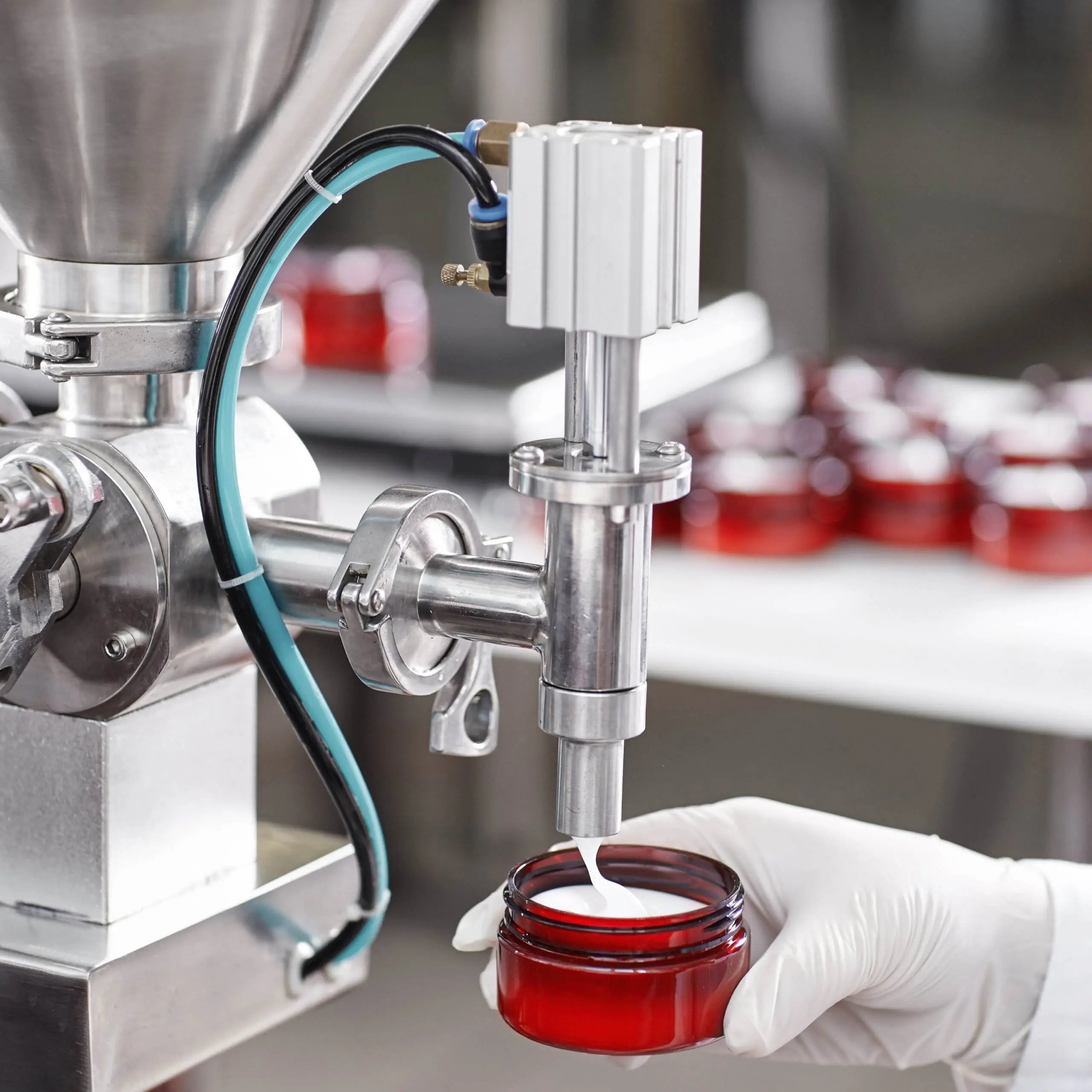 A close-up of a gloved hand filling a jar of face cream underneath a dispenser in a packaging facility.