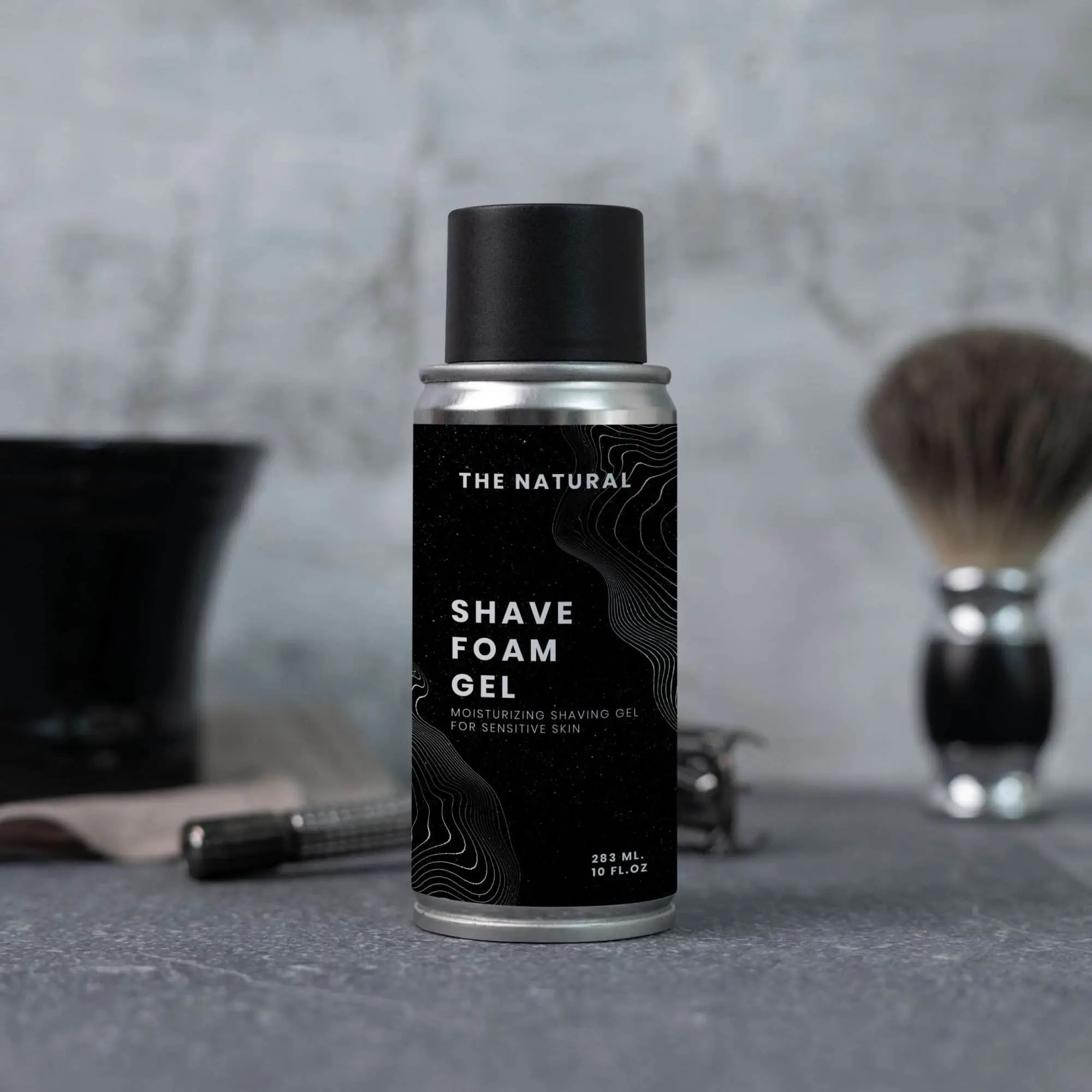 A short aerosol can of shave foam gel with a black label and cap. Behind the can is a safety razor and shave brush.