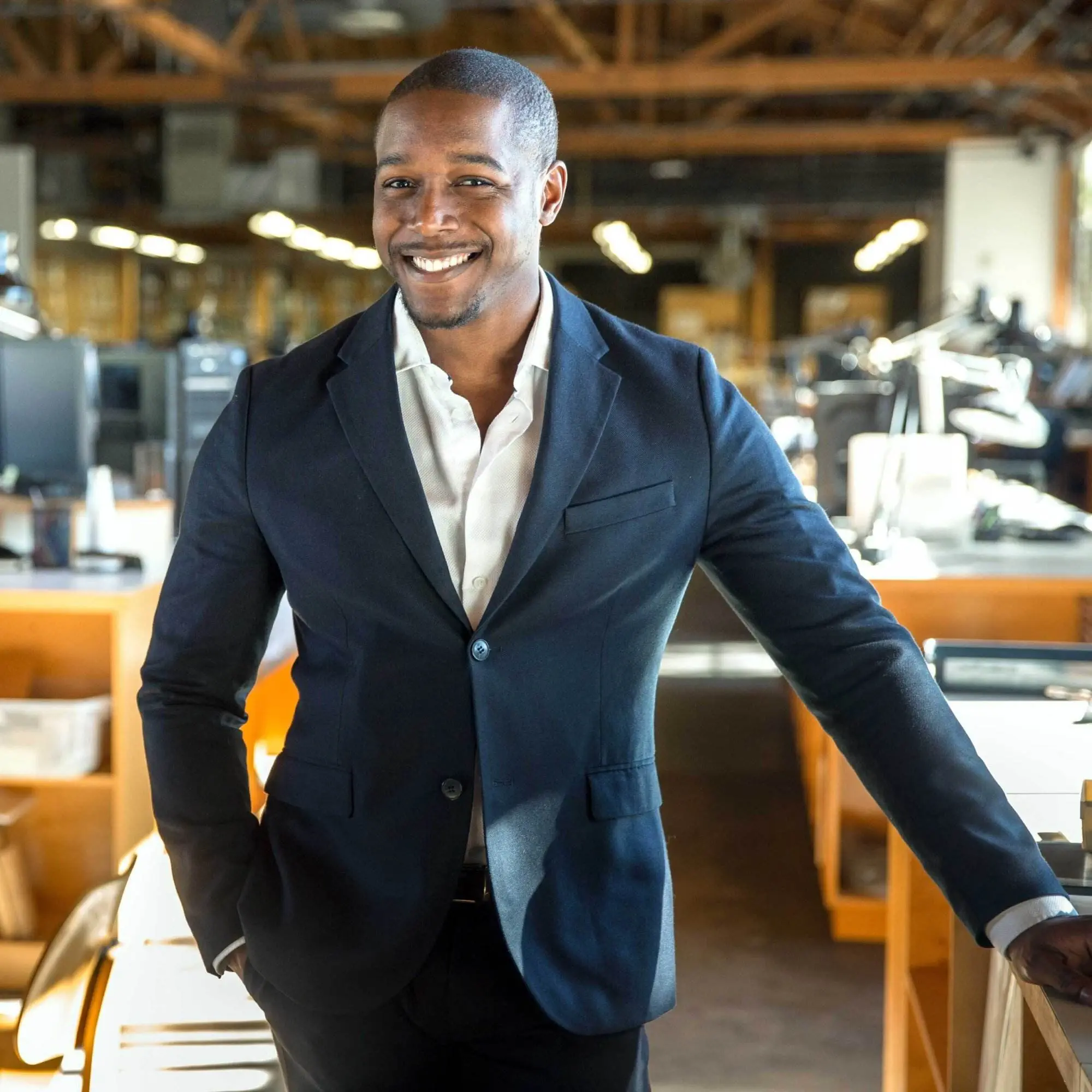 A Black man in a business casual blazer standing in an office. He has his hand on the desk and he's smiling.