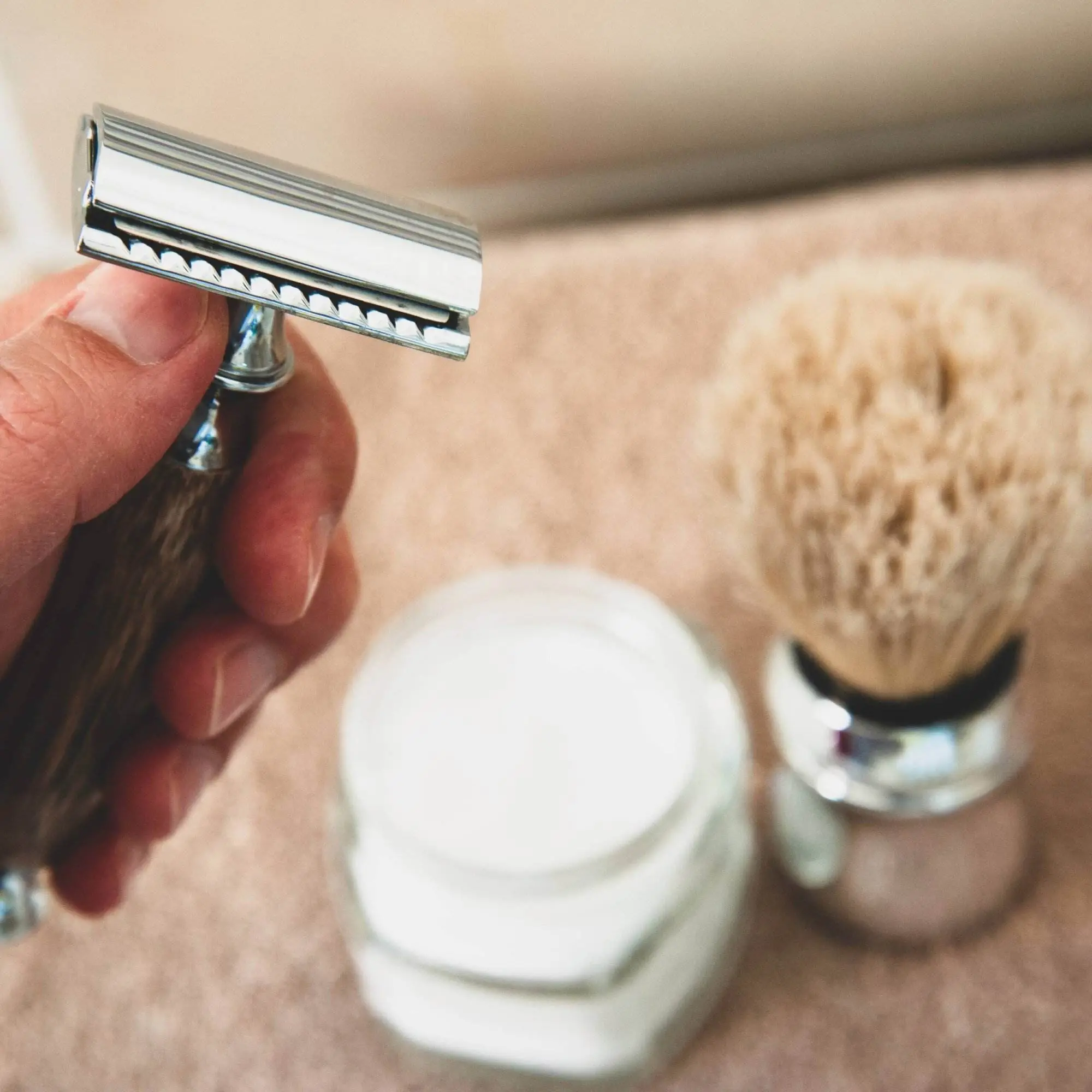 Someone in their bathroom holding a razor near the bathroom vanity, with a shaving brush and shave cream on the counter.