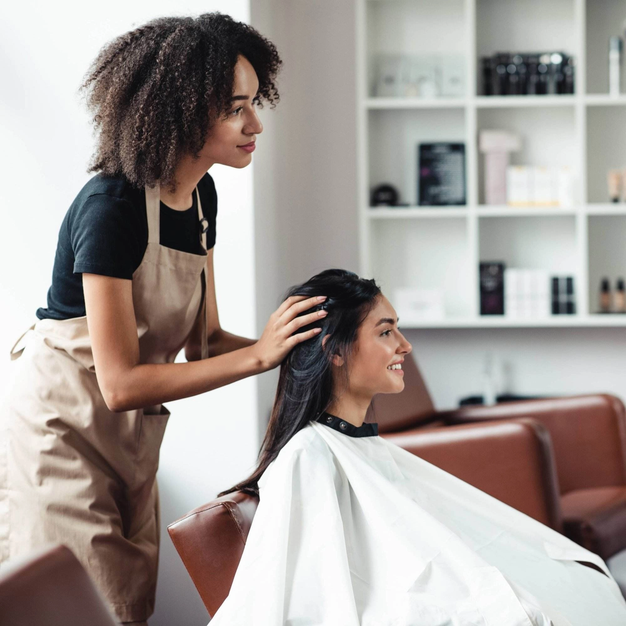 A young female hairstylist assesses a female customer’s hair as she sits in the salon chair, smiling.